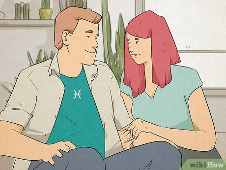 A man sitting on a couch with a woman. Her arms are looped around one of his, and they gaze into each other's eyes. There is a Pisces symbol on his shirt.